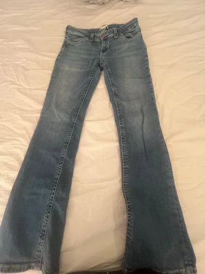 Blå bootcut jeans med kontrastsömmar - Säljer ett par ljusblå bootcut jeans med snygga kontrastsömmar och detaljerade bakfickor. Jeansen har låg midja och klassisk femficksdesign. Perfekta för dig som gillar retrovibbar och vill ha ett par jeans med lite extra attityd.