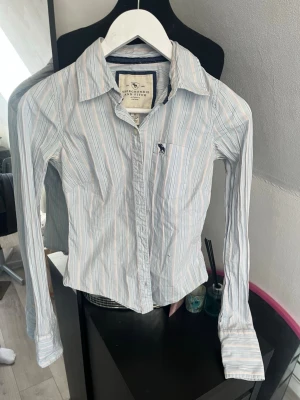 Randig skjorta från Abercrombie - Snygg randig skjorta från Abercrombie & Fitch i ljusblått, vitt och rosa. Skjortan har lång ärm, klassisk krage och knäppning framtill. Broderad logga på bröstfickan. Perfekt för en stilren look.