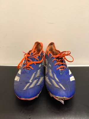 Adidas Predator blå fotbollsskor - Säljer ett par Adidas Predator fotbollsskor i blått med vita detaljer och orange snörning samt häl. Skorna har en låg profil och är designade för bra grepp på planen. Snygg och sportig look som sticker ut på matchen.