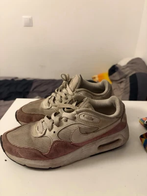 Nike Air Max beige/rosa sneakers - Nike Air Max sneakers i beige och ljusrosa med mesh och mockadetaljer. Klassisk swoosh på sidan och synlig Air-enhet i sulan. Skorna har lågt skaft och snörning framtill. Perfekta för dig som gillar en sportig och stilren look.