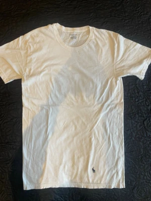 Vit t-shirt från Ralph Lauren - Klassisk vit t-shirt från Ralph Lauren med rund hals och den ikoniska mörkblå broderade polospelaren på nedre delen. Tillverkad i mjuk bomull som känns skön mot huden. Perfekt basplagg för en clean och stilren look.