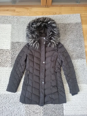 Brun dunjacka från Saki storlek XS - Snygg oanvänd mörkbrun dunjacka med figurnära passform och stor huva med gråsvart fuskpäls. Jackan har dragkedja och knappar framtill samt två fickor med dragkedja. Quiltad design och längre modell som värmer bra under vintern. Storlek XS. Köpt för ca 3000 kr.