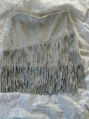 Beige mockakjol med fransar från Zara - Supersnygg grå kortkjol från Zara  i mockaimitation med långa fransar framtill. Kjolen har en dragkedja baktill. Använd som topp🙏🏻 Perfekt för dig som vill sticka ut med en trendig detalj!