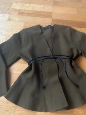 Olivgrön kavaj  - Stilren olivgrön kavaj från med markerad midja och knytband. Kavajen har en modern, något utsvängd passform och är tillverkad i ett mjukt tyg som ger en snygg siluett. Perfekt för dig som vill ha en trendig och unik look.