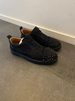 Svarta sneakers med nitar - Snygga mörkblå sneakers från Christian Louboutin i mocka med blå nitar på tån. Skorna har blåa snören, låg profil och en klassisk röd sula på insidan. Perfekta för dig som vill sticka ut med en edgy och exklusiv stil.