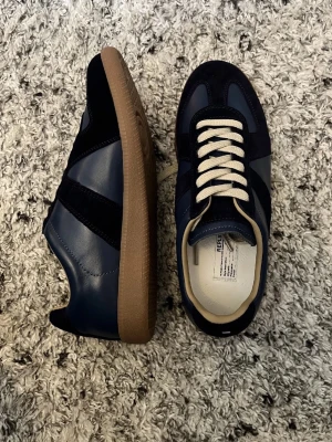  Maison Margiela - Säljer ett par stilrena marinblå Replica sneakers från Maison Margiela gats. Skorna har en kombination av skinn och mocka, vita snören och en klassisk gummisula. Perfekta för dig som gillar en clean och tidlös look. pris kan diskuteras en del 