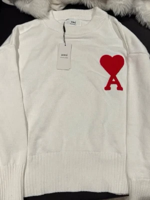 Vit stickad tröja från Ami Paris - Vit stickad tröja från Ami Paris med rund halsringning och ribbade muddar. På bröstet finns ett rött hjärta med bokstaven A, vilket ger en snygg och trendig look. Perfekt för dig som gillar stilrena plagg med en twist. Helt ny säljes då den inte passar