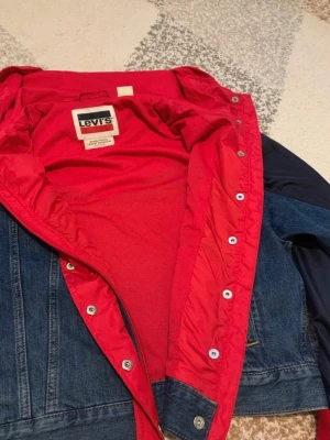 Levi's jeansjacka med röd insida - Snygg jeansjacka från Levi's Premium med klassisk blå denim på utsidan och en cool röd insida. Jackan har tryckknappar framtill och detaljer i kontrastfärg. Perfekt för dig som vill sticka ut med en unik twist på den klassiska jeansjackan.