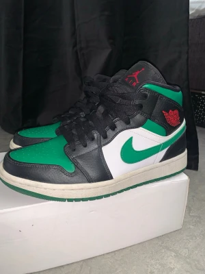 Air Jordan 1 Mid svart/grön/vit - Snygga Air Jordan 1 Mid sneakers i svart, grönt och vitt läder med klassisk röd Jumpman-logga på plösen och röd Wings-logga på sidan. Skorna har svart snörning, vit mellansula och grön yttersula. Perfekta för dig som vill sticka ut med en ikonisk streetwear-look.