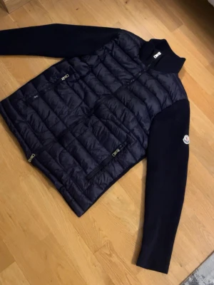 Mörkblå cardiganjacka från Moncler - Snygg mörkblå cardiganjacka från Moncler med quiltad, vadderad front och stickade ärmar. Jackan har dragkedjor framtill och på fickorna samt en hög krage. Moncler-logga på ena ärmen. Perfekt för lager-på-lager och streetstyle.