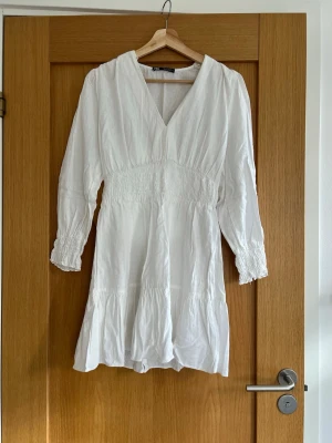 Vit fin kort linne-klänning, med långa ärmar, från Zara - Superfin vit kort klänning från Zara med v-ringning, smock i midjan och långa ärmar med smock vid ärmslut. Tillverkad i ett lätt och luftigt material (50% linne), perfekt för varma dagar och en skolavslutning eller konfirmation🌸🎉! Enkel och stilren design som är lätt att matcha.