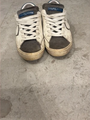 Philippe Model vita och grå sneakers - Snygga sneakers från Philippe Model i vitt läder med grå mockadetaljer på tån och plös samt blå häl. Klassisk låg modell med vita skosnören och gummisula. Perfekta för en avslappnad streetstyle-look.