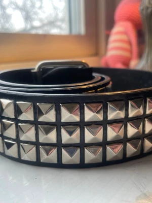 Svart nitarbälte med silverdetaljer - Säljer ett svart bälte med tre rader fyrkantiga silverfärgade nitar. Bältet har en klassisk metallspänne och ger en edgy vibe till vilken outfit som helst. Perfekt accessoar för dig som vill sticka ut lite extra.