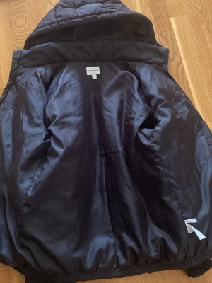 Svart pufferjacka från Kappahl - Säljer en svart pufferjacka från Kappahl med huva och quiltad design. Jackan har en blank insida och diskreta reflexdetaljer. Perfekt för kalla dagar och riktigt skön att bära. Materialet är syntet och jackan har en klassisk pufferlook.