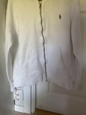 Vit hoodie från Polo Ralph Lauren storlek small - Klassisk vit hoodie från Polo Ralph Lauren med dragkedja framtill och två stora fickor. Liten broderad logga på bröstet. Tillverkad i mjukt bomullsmaterial som känns skönt mot huden. Perfekt för en avslappnad och stilren look. I storlek small. Använd fåtal gånger.