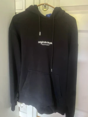 Svart hoodie från Jack & Jones storlek large - Svart hoodie från Jack & Jones Originals med trycket 'Originals Studio' på bröstet. Klassisk känguruficka framtill och justerbar huva med snören. Perfekt för en avslappnad och stilren look. Mjuk bomullsblandning för extra komfort. I storlek large.