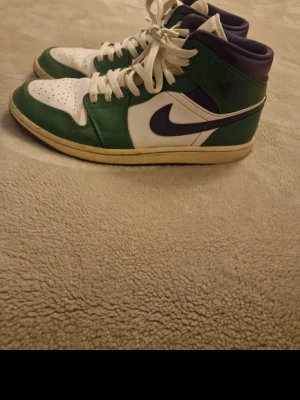 Nike Air Jordan 1 Mid grön/vit/lila - Snygga Nike Air Jordan 1 Mid sneakers i grönt, vitt och lila. Skorna har klassisk hög siluett, vit tåbox med perforeringar, gröna paneler och lila detaljer samt den ikoniska swoosh-loggan på sidan. Ovandelen är i läder och sulan är gulaktig.