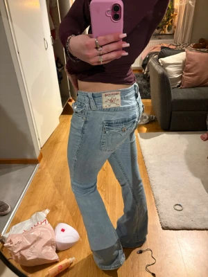 Ljusblå true religion jeans - Säljer ett par ljusblå bootcut jeans med klassisk femficksdesign och låg midja. Jeansen har en avslappnad passform över låren och utsvängda ben. Perfekta för en trendig och avslappnad stil. Säljer då de inte passar mig, mycket bra skick. Skriv för frågor och bilder💕