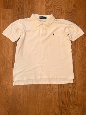 Vit pikétröja från Polo Ralph Lauren - Klassisk vit pikétröja från Polo Ralph Lauren med korta ärmar och krage. Tröjan har knappar framtill och den ikoniska broderade loggan på bröstet. Perfekt för en clean och stilren look.