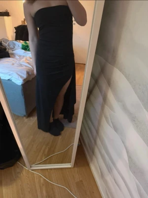 Svart axelbandslös långklänning - Säljer en svart axelbandslös långklänning med en hög slits på ena sidan. Klänningen har en figurnära passform och är perfekt för dig som vill ha en stilren och elegant look. Materialet känns mjukt och följsamt mot kroppen.