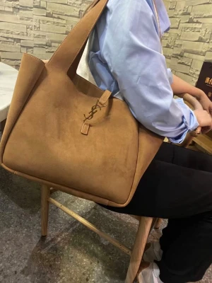 Brun YSL handväska i mocka - Säljer en stor och trendig brun handväska från YSL i mjuk mocka. Väskan har en unik, asymmetrisk form och ett elegant YSL-emblem på framsidan. Perfekt för dig som gillar stilrena och lyxiga accessoarer.