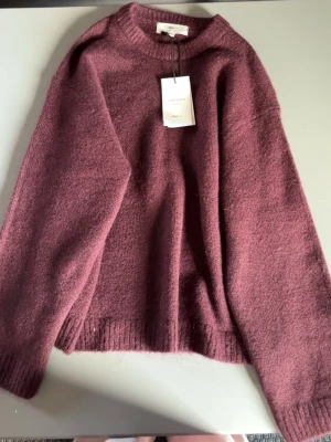 Vinröd stickad tröja från Gina Tricot  - Mysig vinröd stickad tröja  med rund halsringning och ribbade muddar vid ärmslut och nederkant. Tröjan har en avslappnad passform och är tillverkad i ett mjukt, ulligt material som känns skönt mot huden.