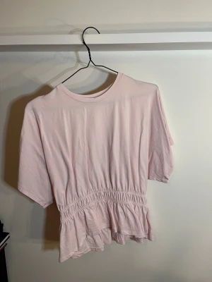 Ljusrosa topp M - Söt ljusrosa topp med korta ärmar och rund halsringning. Toppen har smockad midja som ger en snygg volangdetalj nedtill.