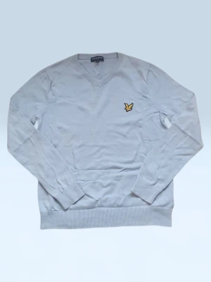 Ljusblå stickad tröja Lyle & Scott - Snygg ljusblå stickad tröja från Lyle & Scott med V-ringning och klassisk gul logga på bröstet. Tröjan har långa ärmar och ribbade muddar vid ärmslut och nederkant. Perfekt för lager på lager och enkel att matcha.