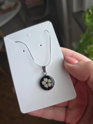 Halsband med blomma i resin - Snyggt halsband med tunn silverfärgad kedja och ett runt hänge i svart resin med en vit torkad blomma inuti. Perfekt accessoar för dig som gillar naturinspirerade smycken och unika detaljer.