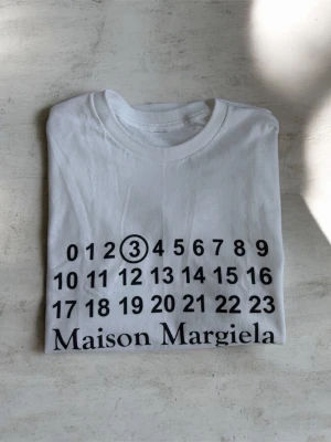 Maison Margiela vit t-shirt - Vit t-shirt från Maison Margiela med ikoniskt tryck av siffror och logga framtill. Klassisk rund hals och korta ärmar. Perfekt för dig som gillar minimalistisk och stilren design. Vid snabb affär kan pris diskuteras 
