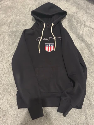 Gant hoodie  - Svart hoodie från GANT med vit dragsko i huvan och stor broderad logga samt amerikanskinspirerat sköldmärke på bröstet. Klassisk känguruficka framtill och ribbade muddar. Perfekt för en avslappnad och stilren look.