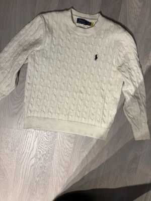 Vit kabelstickad tröja Polo Ralph Lauren - Kabelstickad vit tröja från Polo Ralph Lauren med rund halsringning och den klassiska broderade loggan på bröstet. Tröjan har ribbade muddar vid ärmslut och nederkant och är tillverkad i mjukt material som känns lyxigt. Passar folk runt 170-185cm