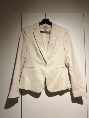 Beige kavaj från H&M, storlek 36 - Stilren beige kavaj från H&M med klassisk passform och en knapp framtill. Kavajen har smala slag, två fickor och är helfodrad. Perfekt för en snygg och uppklädd look. Materialet känns mjukt och har en lätt glans.