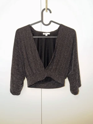 Glittrig svart croppad omlottopp - Snygg svart croppad omlottopp från Gina Tricot med glittrigt material. Toppen har trekvartsärm och djup v-ringning framtill. Perfekt för att sticka ut med en trendig look.