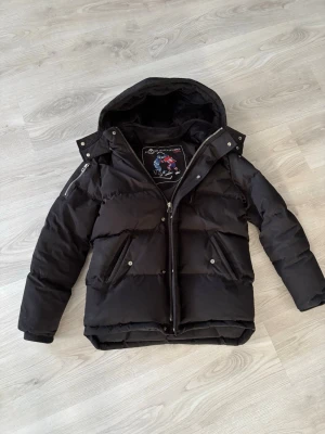 Svart pufferjacka från Moose Knuckles - Säljer en svart pufferjacka från Ice Knockers med huva och dragkedja. Jackan har två stora fickor med tryckknappar och en extra ficka på ärmen. Perfekt för kalla vinterdagar och riktigt skön att bära. Materialet är slitstarkt och håller värmen bra. Pris kan diskuteras