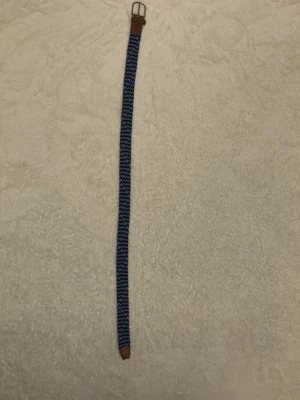 Blått flätat bälte med läderdetaljer - Snyggt flätat bälte i marinblått och ljusblått med bruna läderdetaljer vid spännet och änden. Bältet har ett randigt mönster och passar perfekt till jeans eller chinos för en avslappnad men stilren look.