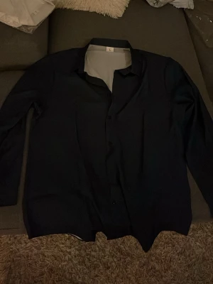 Mörkblå skjorta i klassisk modell - Snygg mörkblå skjorta med klassisk krage och knäppning framtill. Skjortan har lång ärm och en stilren, enkel design som passar till många olika outfits. Perfekt för dig som gillar en clean och tidlös look.