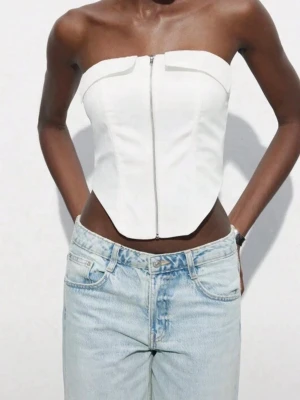 Vit korsett-topp med dragkedja - Säljer en snygg vit korsett-topp med dragkedja framtill. Toppen är axelbandslös och har en strukturerad passform som framhäver midjan. Perfekt att styla med jeans eller kjol för en trendig look. i nyskick aldrig använt