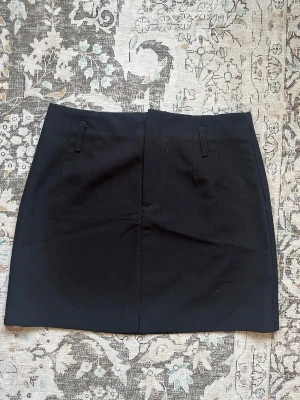 Svart kortkjol  - En stilren svart kortkjol med enkel design och bälteshällor i midjan. Kjolen har rak passform och är tillverkad i ett kostym material som passar perfekt till olika outfits. Inga synliga detaljer förutom en liten slit fram för komfort, klassisk och clean look.