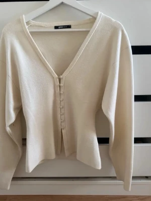 Krämvit kofta från Gina Tricot - Säljer en krämvit kofta från Gina Tricot med v-ringning och dekorativa knappar framtill. Koftan har lång ärm och är tillverkad i ett mjukt, tunt stickat material. Enkel och stilren design som passar till mycket.