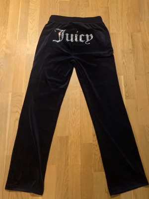 Juicy Couture byxor  - Mörkblåa velour-mjukisbyxor från Juicy Couture med glittrande 'Juicy'-text baktill och broderad logga fram. Byxorna har raka ben, resår och dragsko i midjan för skön passform. Perfekta för en avslappnad och trendig look.