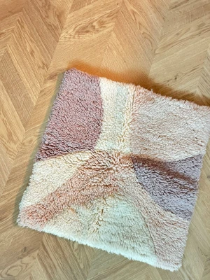 Kuddfodral i pastellfärger - Säljer ett fyrkantigt kuddfodral med mjuk, fluffig framsida i pastelliga toner av beige, rosa och lila. Baksidan är i slät, ljus textil med öppning för kudde. 50*50cm