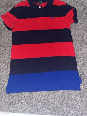 Randig pikéskjorta från Polo Ralph Lauren - Randig pikéskjorta från Polo Ralph Lauren i rött, marinblått och blått. Skjortan har korta ärmar, klassisk krage med knappar och broderad logga på bröstet. Tillverkad i mjuk bomull för en bekväm känsla.