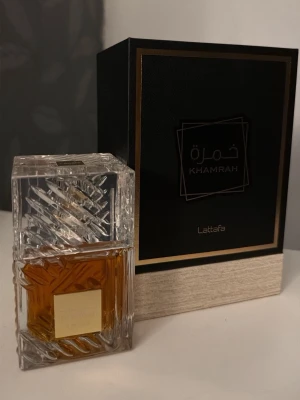 Lattafa Khamrah parfym 100ml - Lattafa Khamrah parfym i en lyxig, rektangulär glasflaska med dekorativa mönster och guldig etikett. Parfymen har en varm bärnstensfärgad vätska och levereras i en elegant svart och beige kartong med gulddetaljer.