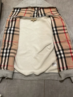 Grå jacka med Burberry-rutor - Snygg grå jacka från Burberry med det klassiska rutiga fodret i beige, svart, rött och vitt. Jackan har dragkedja och är perfekt för dig som vill ha en stilren look med lyxig känsla. Ikoniskt mönster på insidan och mjukt material.