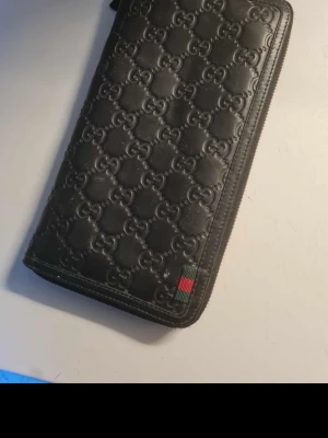 Svart Gucci plånbok i läder - Snygg svart plånbok från Gucci i äkta läder med präglat GG-mönster över hela ytan. Plånboken har dragkedja runtom och en liten detalj med grönt och rött band. Perfekt för dig som gillar exklusiva accessoarer och vill ha plats för både kort och sedlar.