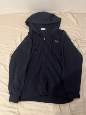 Mörkblå hoodie från Lacoste - Snygg mörkblå hoodie från Lacoste med dragkedja framtill och klassisk krokodillogga på bröstet. Hoodien har huva med dragsko och ribbade muddar vid ärmslut och nederkant. Perfekt för en chill och stilren look. Hoodien är XL men passar även till folk som har L, och så är den regular fit som betyder att den inte är så loose och Inte heller så tight!