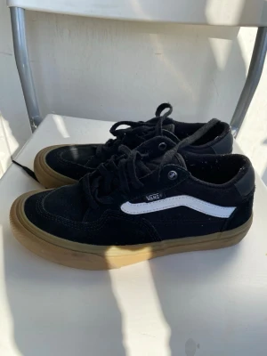 Svarta Vans sneakers med gummisula - Svarta Vans sneakers med klassisk vit sidestripe och brun gummisula. Skorna har lågt skaft, svart canvas och mockadetaljer samt snörning framtill. Perfekta för en streetwear-look och passar till det mesta.