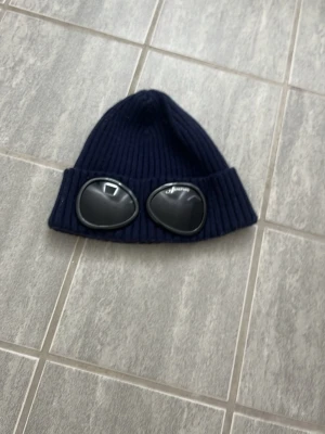 Mörkblå C.P. Company goggle mössa - Säljer en mörkblå ribbstickad mössa från C.P. Company med de ikoniska svarta goggle-linserna framtill. Mössan är tillverkad i mjukt, stretchigt material och har en uppvikt kant. Perfekt för dig som vill ha en streetwear-inspirerad look.