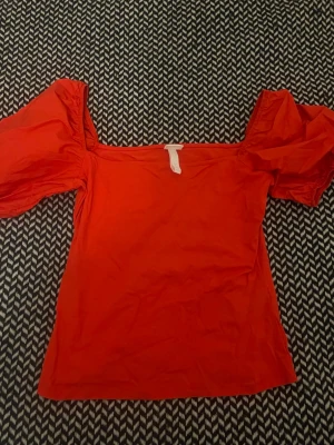 Röd topp med puffärmar - Trendig röd topp med stora puffärmar och fyrkantig urringning. Toppen är axelbandslös och har en figurnära passform som ger en snygg siluett. Perfekt för dig som vill sticka ut med en färgstark och modern look.